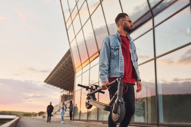 Bel giovane ragazzo in abiti casual in piedi con lo scooter elettrico all'aperto durante il giorno soleggiato vicino all'edificio degli affari