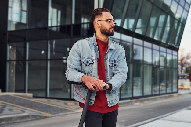 Bel giovane ragazzo in abiti casual in piedi con lo scooter elettrico all'aperto durante il giorno soleggiato vicino all'edificio degli affari