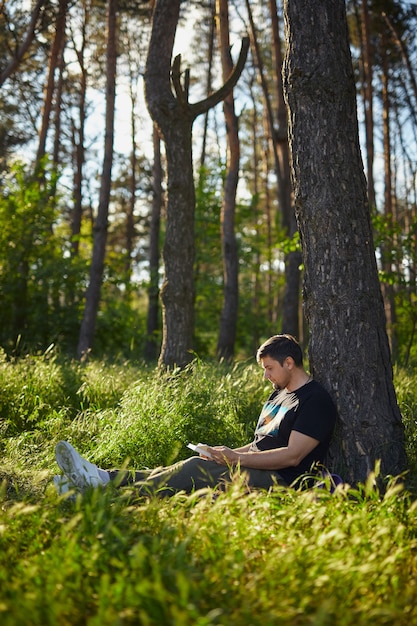 Bel giovane seduto nella foresta sull'erba, appoggiato a un albero, leggendo un libro