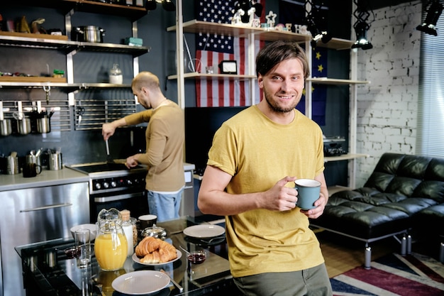 Bel giovane sorridente attraente che beve una tazza di caffè mattutino quando il suo ragazzo cucina la colazione in background