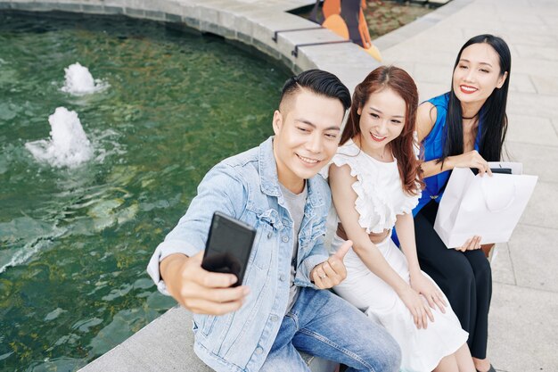 Bel giovane uomo asiatico seduto alla fontana con i suoi amici femminili e prendendo selfie dopo lo shopping insieme nel centro commerciale