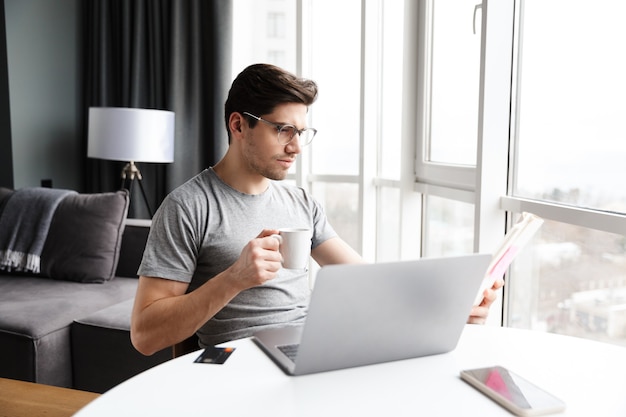 Bel giovane uomo barbuto che indossa abiti casual utilizzando il computer portatile mentre è seduto al tavolo a casa, bevendo caffè