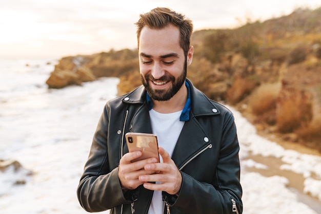 Bel giovane uomo barbuto che indossa una giacca usando il telefono cellulare mentre si trova in spiaggia