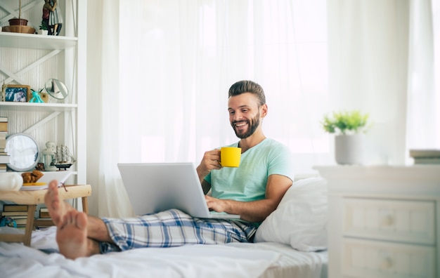 Bel giovane uomo barbuto in abiti casual sta lavorando a casa mentre era seduto sul letto. Ragazzo fiducioso con un laptop e uno smartphone beve caffè in camera da letto.