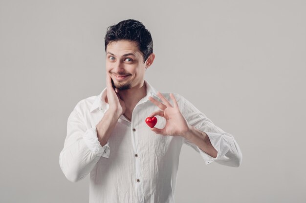 Bel giovane uomo bruna in camicia bianca tiene in mano un cuore rosso su sfondo grigio.