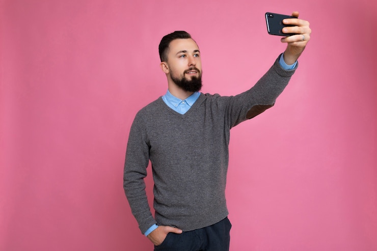 Foto bel giovane uomo che indossa abiti casual alla moda in piedi isolato su sfondo muro tenendo lo smartphone prendendo selfie foto guardando il display del telefono cellulare