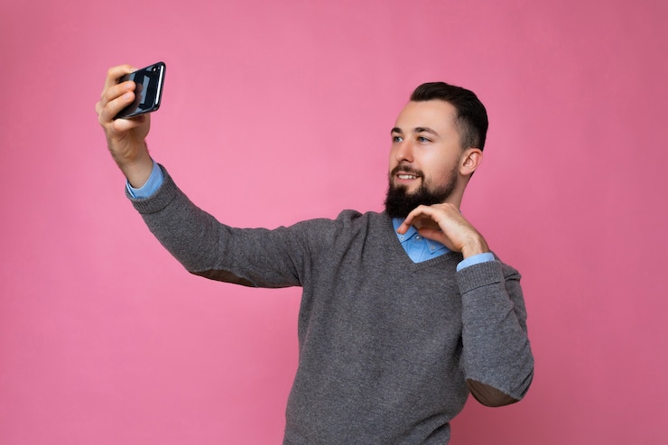 Foto bel giovane uomo che indossa abiti casual alla moda in piedi isolato su sfondo muro tenendo lo smartphone prendendo selfie foto guardando il display del telefono cellulare