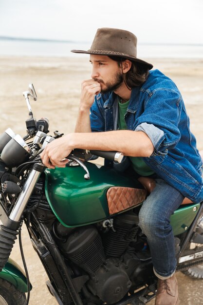 Bel giovane uomo che indossa abiti casual seduto su una moto in spiaggia, indossando un cappello