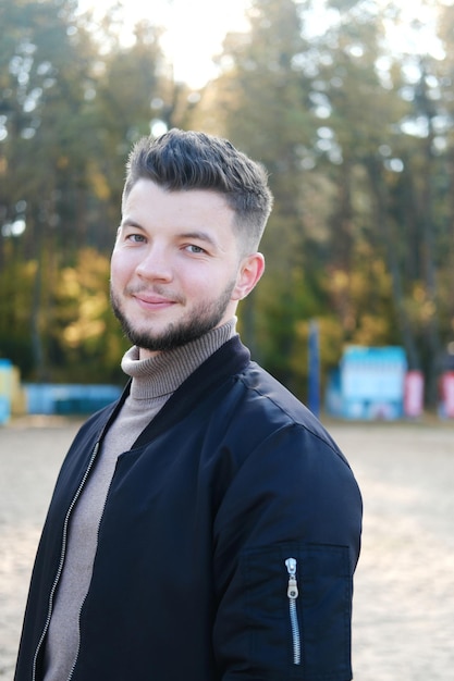 Bel giovane uomo con barba e in una giacca nera in posa sulla spiaggia L'uomo sorride e guarda la macchina fotografica