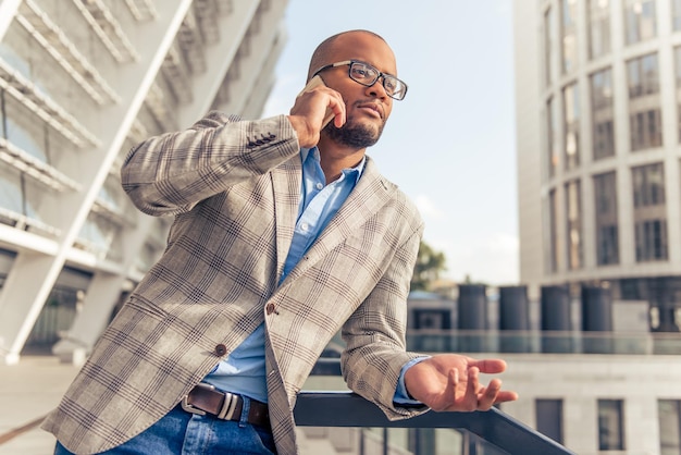 Bel giovane uomo d'affari afroamericano con gli occhiali sta parlando al telefono cellulare in piedi all'aperto e appoggiato sulla balaustra del balcone