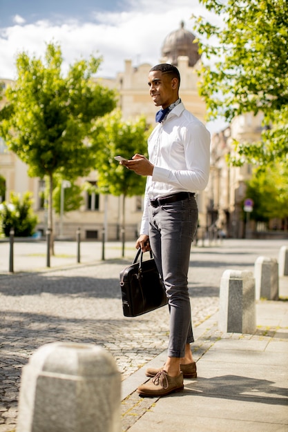 Bel giovane uomo d'affari afroamericano utilizzando un telefono cellulare mentre waitng per un taxi su una strada