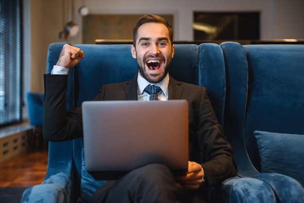 Bel giovane uomo d'affari che indossa tuta seduto nella hall dell'hotel, utilizzando il computer portatile, celebrando