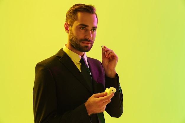 Bel giovane uomo d'affari che indossa un abito formale in piedi isolato su un muro giallo, con in mano una scatola con auricolari wireless