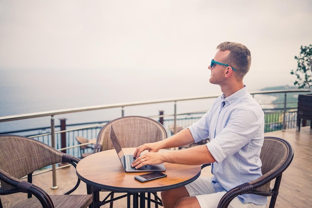 Bel giovane uomo d'affari di successo seduto a un tavolo a bordo piscina con un laptop con vista sul Mar Mediterraneo Lavoro a distanza in vacanza Concetto di vacanza