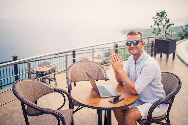 Bel giovane uomo d'affari di successo seduto a un tavolo a bordo piscina con un laptop con vista sul Mar Mediterraneo Lavoro a distanza in vacanza Concetto di vacanza