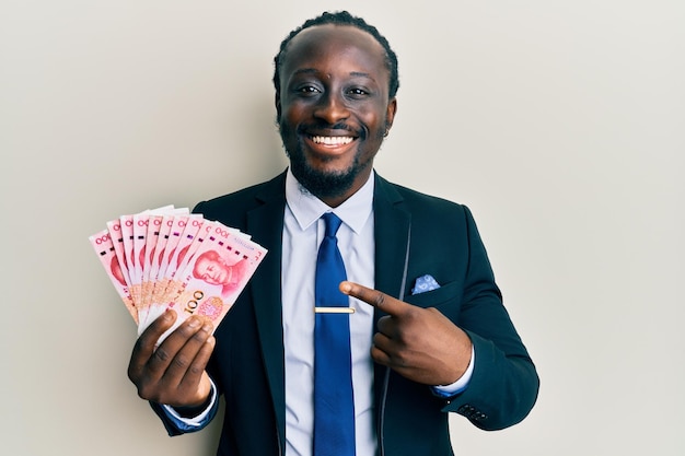 Bel giovane uomo di colore che indossa un completo da lavoro in possesso di banconote da 100 yuan sorridendo felice indicando con la mano e il dito