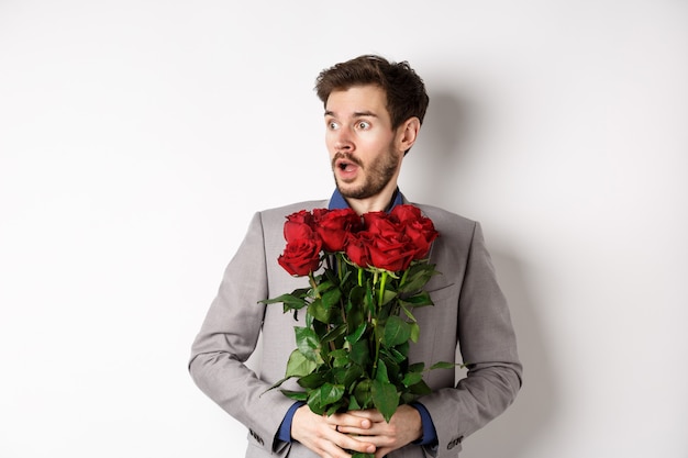 Bel giovane uomo in tuta in possesso di rose rosse, guardando a sinistra con espressione sorpresa e allarmata, in piedi il giorno di San Valentino su sfondo bianco.