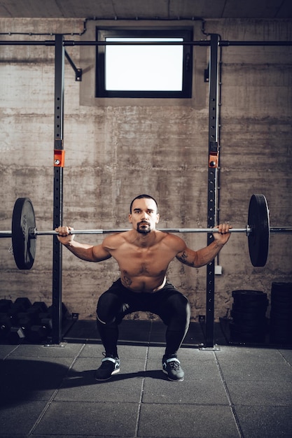 Bel giovane uomo muscolare che fa esercizio tozzo con bilanciere in palestra. Guardando la fotocamera.