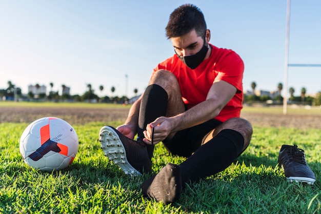 Bel giovane uomo seduto con la maschera covid19 che allaccia i lacci delle scarpe da calcio