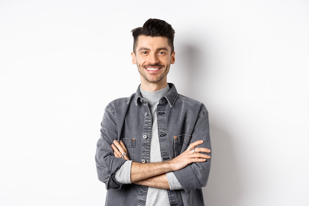 Bel giovane uomo sorridente con le braccia incrociate sul petto, sentendosi pronto, guardando motivato alla telecamera, in piedi su sfondo bianco.