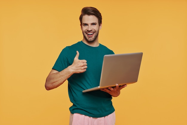 Bel giovane uomo sorridente in abbigliamento casual che trasporta laptop e gesticola mentre si trova in piedi contro il muro giallo