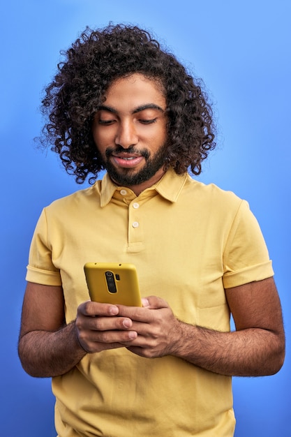 Bel maschio arabo utilizza lo smartphone, chiacchierando con un amico, digitando un messaggio. isolato su muro blu