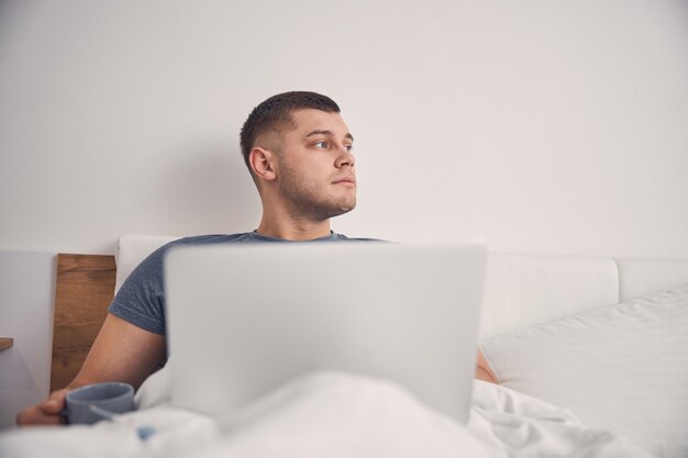 Bel maschio bruna sdraiato a letto con il computer mentre tiene in mano una tazza di caffè
