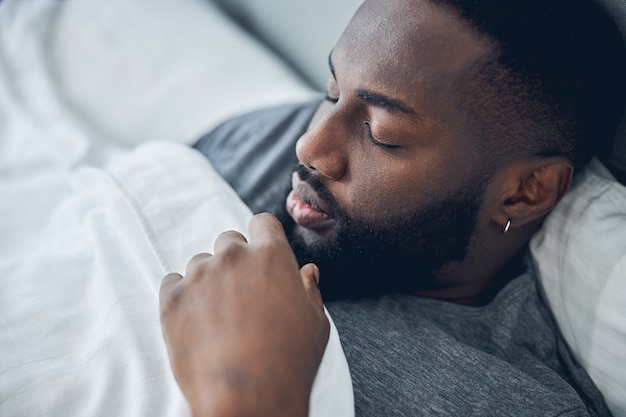 Bel maschio dalla pelle scura che dorme al mattino mentre giace sotto una coperta calda