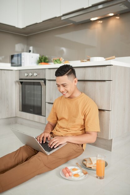 Bel maschio seduto sul pavimento e tenendo il computer portatile sulle ginocchia con la sua colazione.