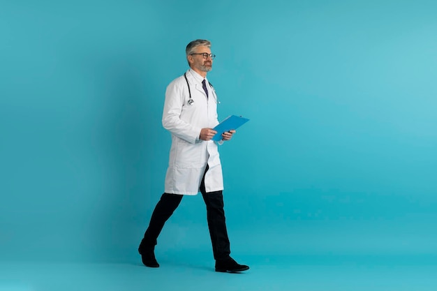 Bel medico di mezza età con cartella medica che cammina sul blu