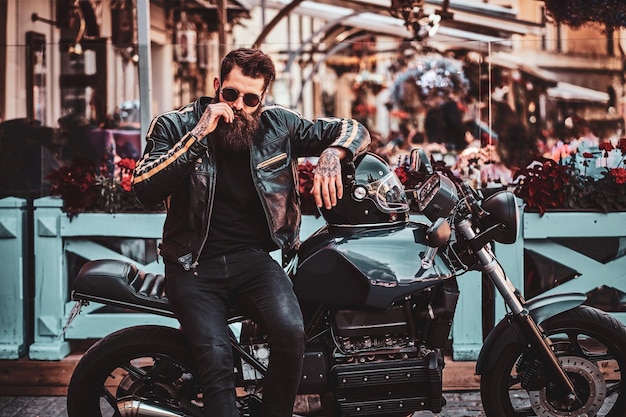 Bel motociclista serio con barba e tatuaggi è seduto sulla sua moto nel vivace centro cittadino.