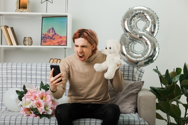 Bel ragazzo arrabbiato durante la giornata delle donne felici che tiene orsacchiotto guardando il telefono in mano seduto sul divano nel soggiorno