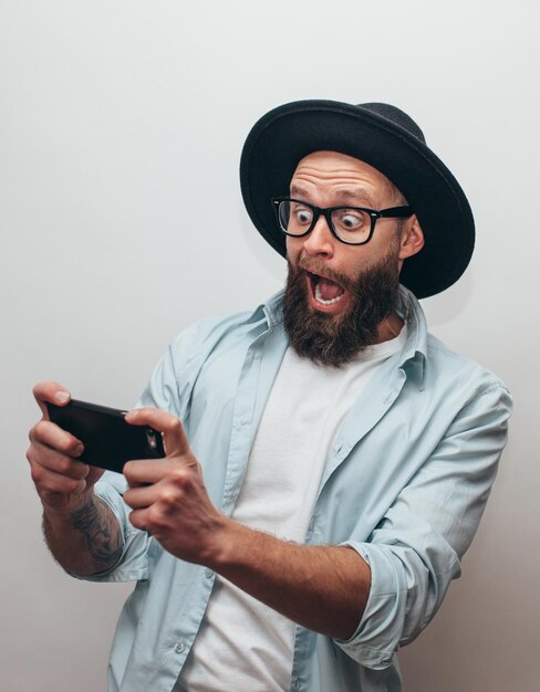 Bel ragazzo barbuto tiene smartphone e gioca online Uomo gioioso che indossa un cappello nero elegante e una camicia blu Emozioni pazze