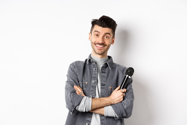 Bel ragazzo caucasico con i baffi incrociate le braccia sul petto, tenendo il microfono e sorridendo alla telecamera, esibirsi sul palco con il microfono, in piedi su sfondo bianco.