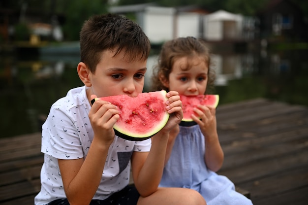 Bel ragazzo che mangia una fetta di deliziosa anguria dolce e succosa, seduto accanto a sua sorella minore. Godendo la giornata estiva sul molo di campagna.