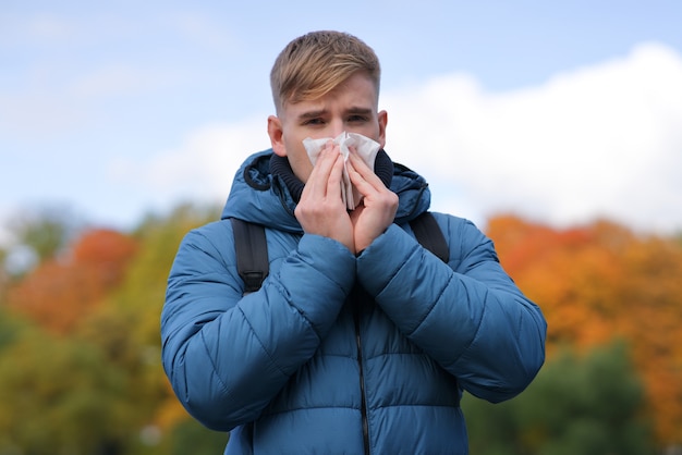 Bel ragazzo che si soffia il naso in un fazzoletto di carta all'aperto su uno sfondo autunnale dorato naturale