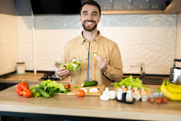 Bel ragazzo con la barba e un bel sorriso sta cucinando insalata. Probabilmente gli piace uno stile di vita sano