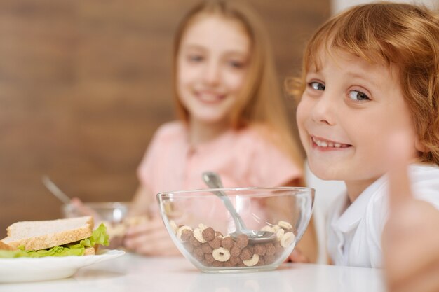 Bel ragazzo energico sincero seduto al tavolo e mangiare cereali dolci mentre mostra quanto gli piace