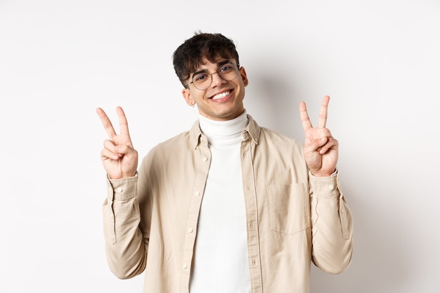 Bel ragazzo hipster con gli occhiali, sorridente con i denti bianchi, che mostra un gesto di pace o vittoria, in piedi ottimista su sfondo bianco