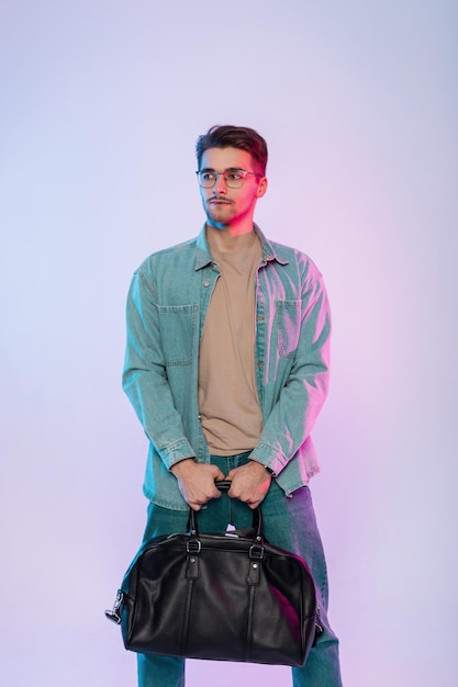 Bel ragazzo hipster uomo d'affari con acconciatura e occhiali vintage in abito casual in denim con jeans e borsa in pelle si trova in studio con luci colorate rosa e blu