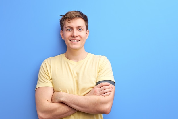 Bel ragazzo in maglietta casual sorridendo alla telecamera isolato su sfondo blu, giovane maschio in piedi con le braccia conserte, incrociate. persone, concetto di stile di vita