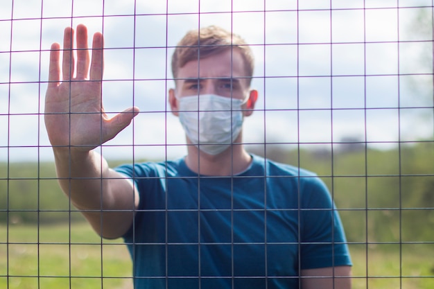 Bel ragazzo in maschera protettiva sul viso in piedi dietro le sbarre, reticolo. Limitazione della libertà. Autoisolamento, infezione da virus dell'epidemia di coronavirus. Covid19. Palma isolata di manifestazione del giovane, fanale di arresto