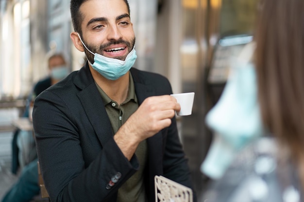 Bel ragazzo in tuta con maschera facciale abbassata che parla e beve caffè espresso in un caffè all'aperto