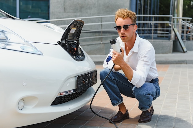 Bel ragazzo seduto vicino alla sua nuova auto elettrica moderna e tenendo la spina del caricabatterie, mentre l'auto si sta caricando alla stazione di ricarica