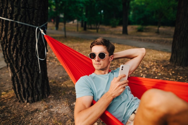 Bel ragazzo serio in occhiali da sole e abiti casual giace su un'amaca su un albero nel bosco