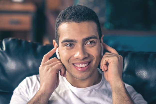 Bel ragazzo sorridente che ascolta la musica in cuffie wireless bianche, toccandole con le mani.