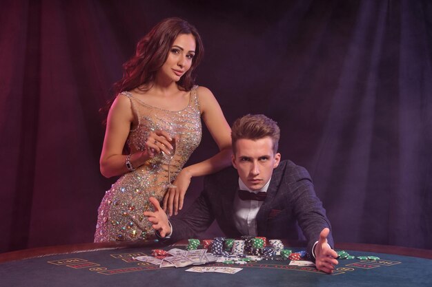 Bel ragazzo sta giocando a poker al casinò al tavolo con pile di fiches e carte su di esso Egli celebra la vittoria con una splendida femmina nero fumo sfondo retroilluminazione colorata gioco d'azzardo primo piano