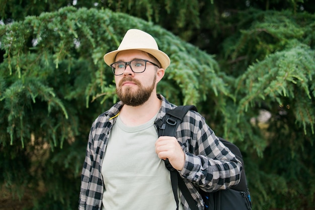 Bel ragazzo turistico con zaino che indossa un cappello di paglia per viaggiare in piedi contro la schiena dell'albero verde