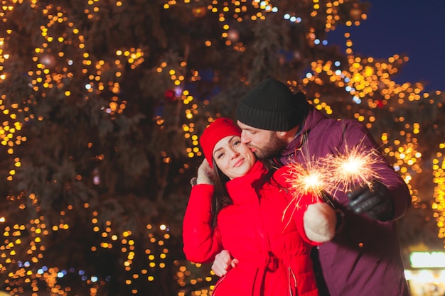 Bel ritratto di giovane uomo bello che bacia la guancia di sua moglie, tenendole la vita, entrambi illuminando scintille. Grande albero di Natale all'aperto con luci sullo sfondo