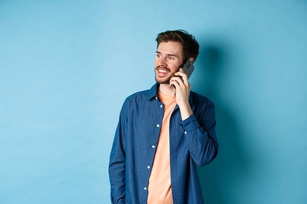 Bel uomo caucasico parlando al telefono cellulare, guardando a parte lo spazio vuoto con un sorriso spensierato, in piedi su sfondo blu.